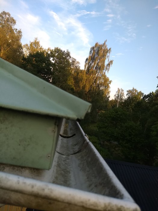 Öppnad hängränna med uppåtvikt plåt för att möjliggöra åtkomst till skruvar, med skymt av träd i bakgrunden.