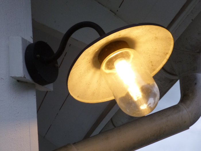 Utomhusvägglampa med tänd glödlampa under taknock, tillgänglig på Jula och Clas Ohlson.