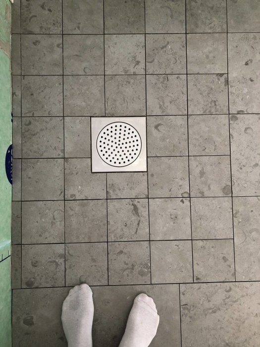 Fötter i vita strumpor står på nykapad granitkeramikgolv nära en brunnslock, redo för fogning.