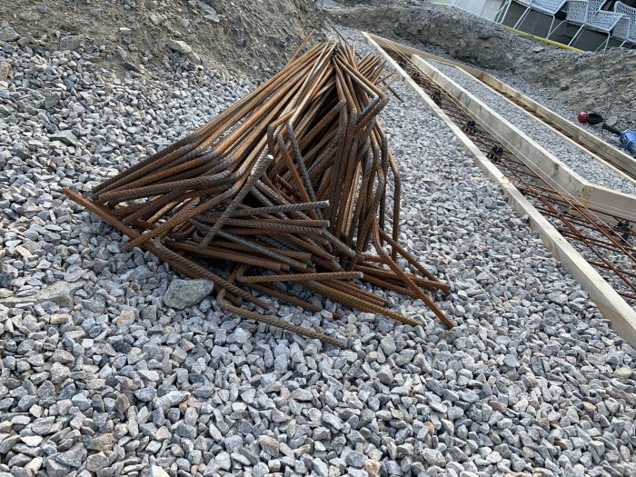 Rostiga armeringsjärn i hög på makadam med träformar för gjutning av fundament i bakgrunden.