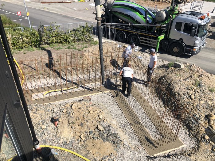 Byggplats med armeringsjärn, betongbil och personer som förbereder för gjutning av fundament.