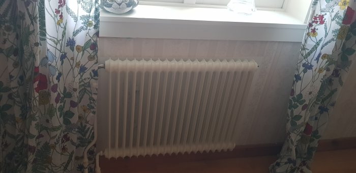 Vit radiator under ett fönster med ljust tapetserade väggar och blommiga gardiner.