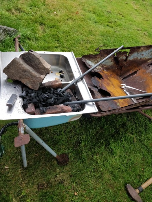 Rostig, sönderfallande grill med kol och stenar i gräsmiljö, verktyg syns på marken.