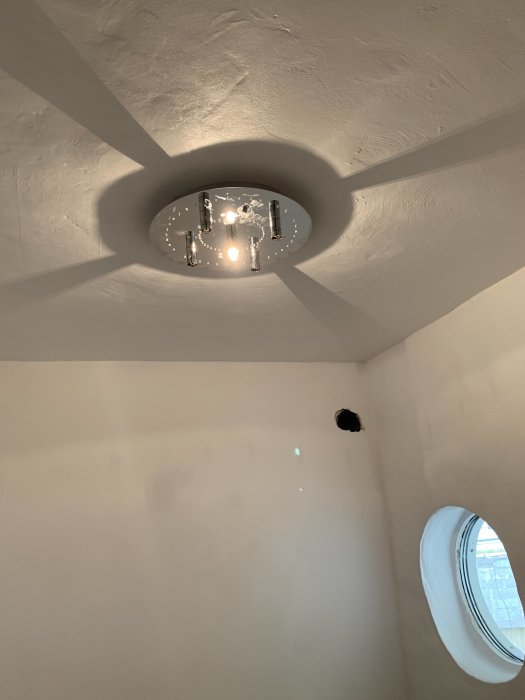 Tak med öppen ljuskälla och synliga ledningar samt ett runt fönster på väggen.