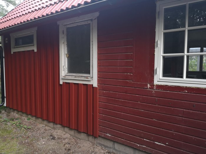 Gammal rödmålad stuga med kombination av stående och liggande träpanel, samt två fönster i olika stilar.