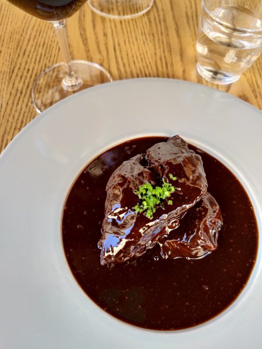En tallrik med oxkind serverad i en mörk sås, prydd med grön garnering, med ett glas rött vin i bakgrunden.