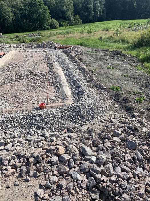 En grävd dräneringsdike fylld med makadam med synligt fall riktat mot en samlingsbrunn i en trädgård.