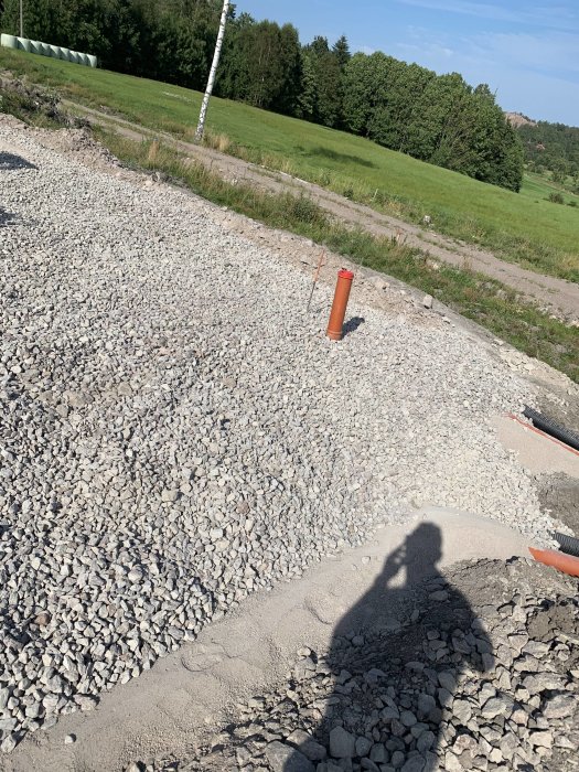 Grusmark med utstickande rörbitar för dagvattenanslutning, skugga av fotografen, grönt landskap i bakgrunden.