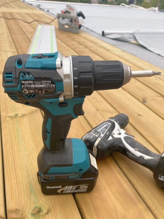 Makita DDF 484 skruvdragare på trämaterial med kapsåg i bakgrunden.