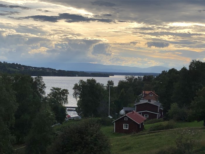 Vy över en sjö med en röd stuga i förgrunden och dramatisk himmel med moln ovanför.