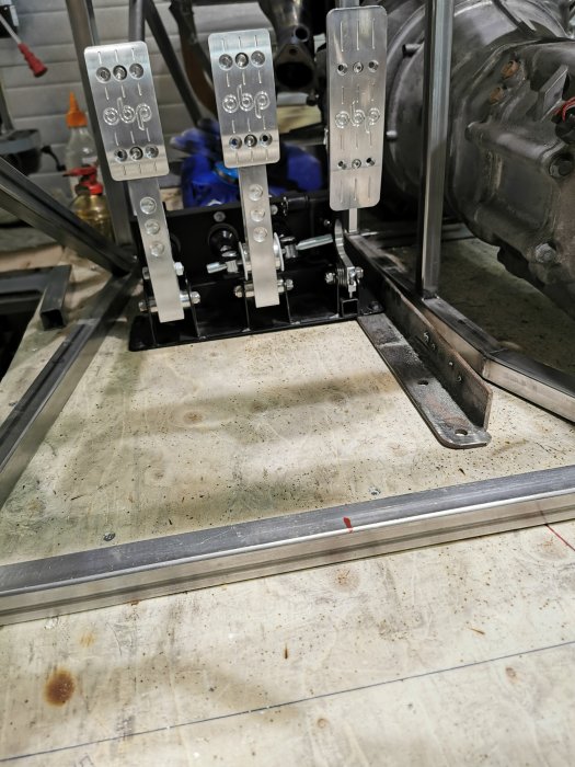 Pedalställ med tre metallpedaler monterat i en fordonskonstruktion, omgivet av stålrör och vinkeljärn.