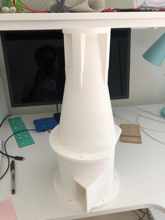 3D-utskriven vit konisk objekt, likt en dammsugardel, på ett skrivbord med en datorskärm i bakgrunden.