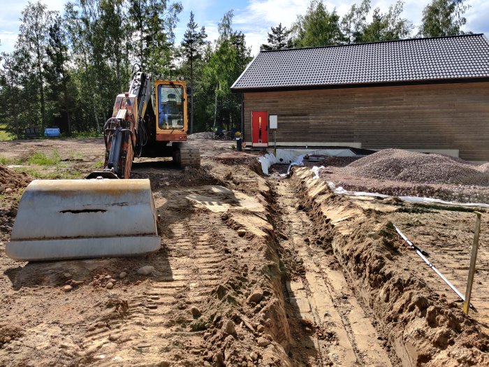 Grävmaskin vid grävd grund till hus med grushögar och byggmaterial i bakgrunden.