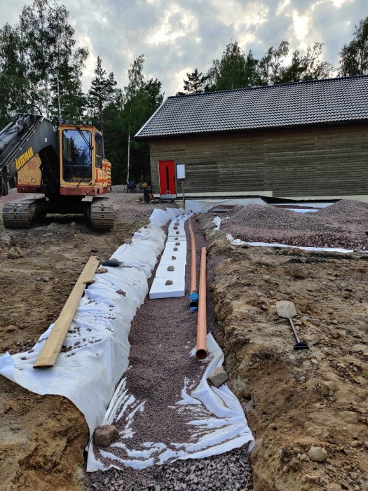 Grävd grund med isolering och rörledningar vid byggplats, grävmaskin i bakgrunden.