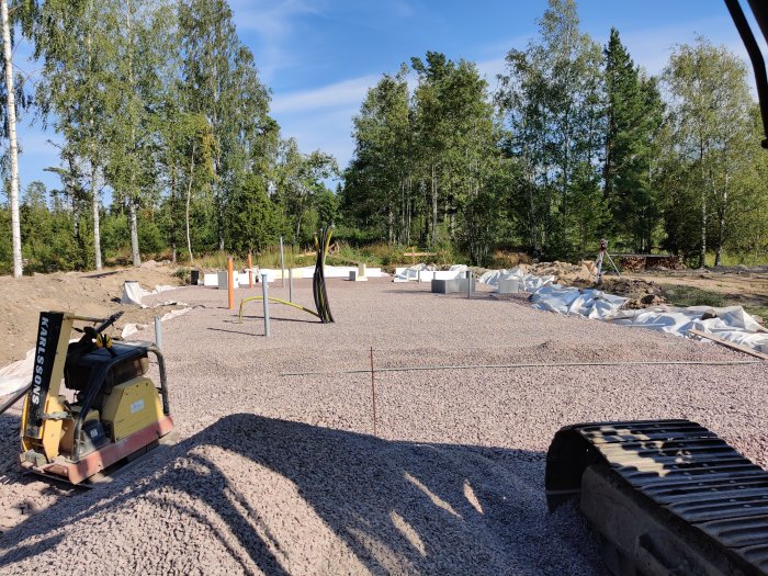 Byggarbetsplats med pågående grundarbete, kantelement och isoleringsmaterial, och en kompaktor i förgrunden.