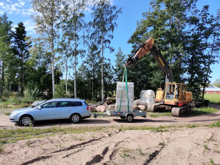 Byggplats med grävmaskin som lyfter byggmaterial bredvid en bil med släp på en grusig yta med träd i bakgrunden.