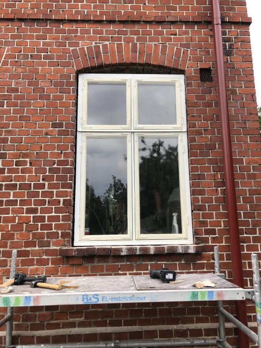 Renoverat vitt fönster på rödtegelbyggnad, verktyg på byggställning framför.