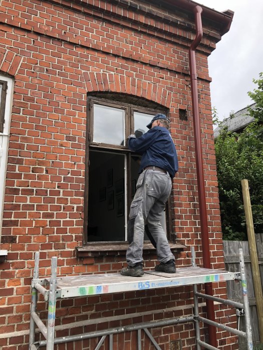 Person renoverar ett gammalt fönster på en tegelbyggnads bottenvåning, står på byggställning.