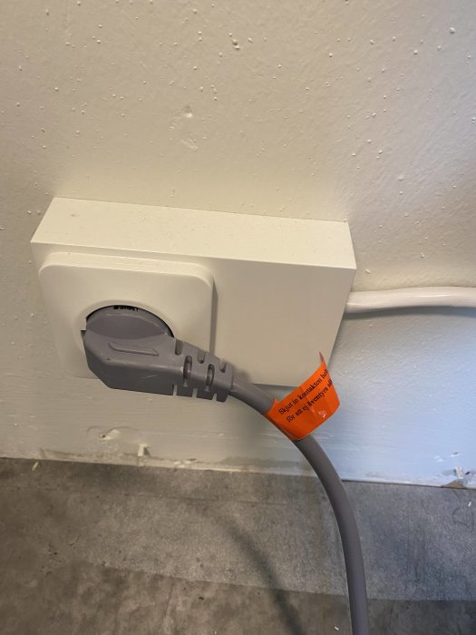 Perilexuttag med en inkopplad sladd och ett synligt varningsetikett på kabeln mot en vägg med synliga märken.