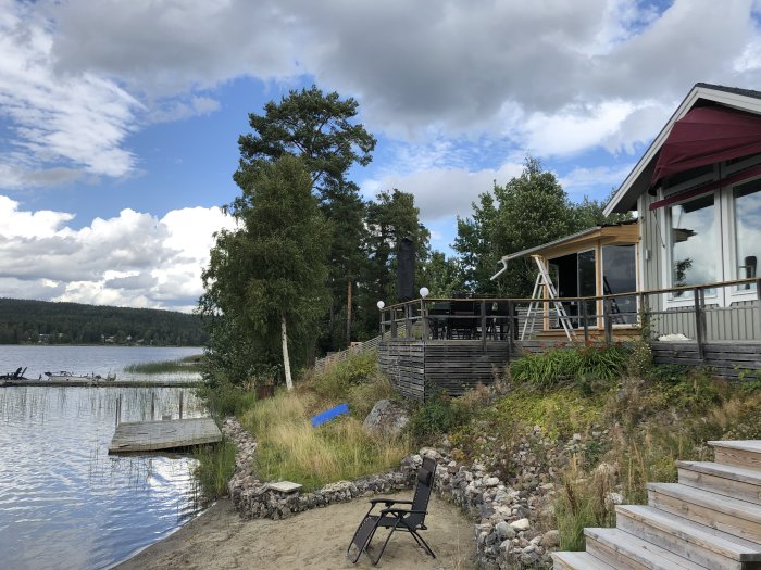 Hus med stor terrass intill en sjö med en brygga och sandstrand, omgivet av träd och vegetation under molnig himmel.