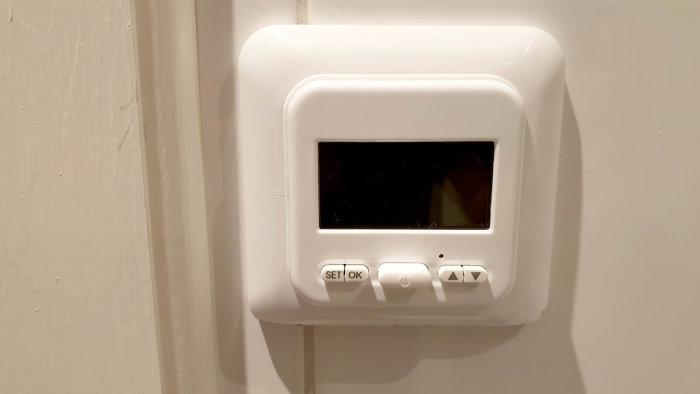 Vit väggmonterad termostat utan synlig märkning eller produktnamn, med bildskärm och knappar.