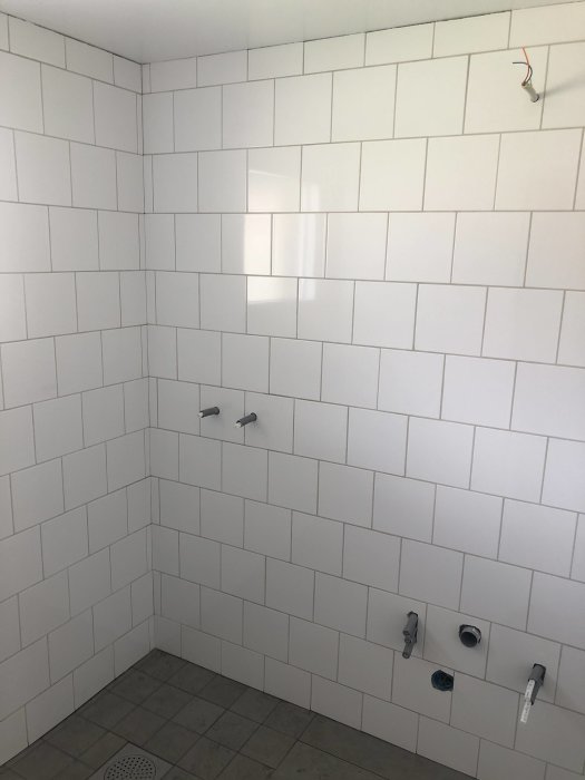 Nytt kaklat badrum med vita väggar och grått klinkergolv med installationsrör synliga.