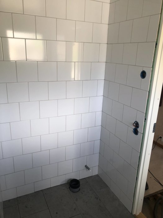Nyligen fogat badrum med vita kakelväggar och grå klinkergolv med installationsdetaljer synliga.