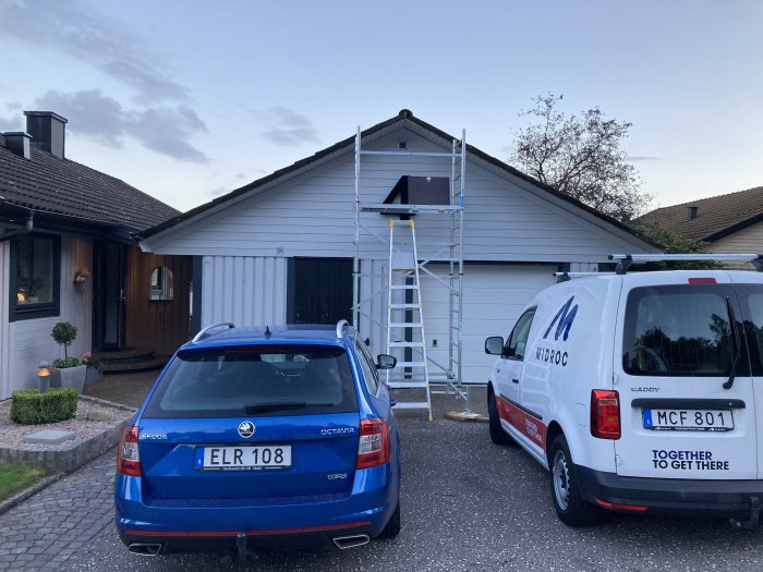 Renoveringsarbete på hus med ställning framför taket och två parkerade bilar, en blå och en vit firmabil.