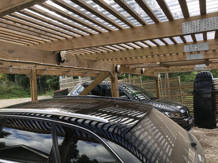 Carport med tak av träribbor, bilar parkerade, och lampor monterade på stolpar.