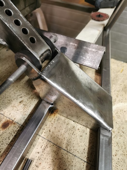 Metallkonstruktion som hålls i en skruvstycke, synliga svetsfogar och bearbetade metallytor indikerar pågående byggarbete.