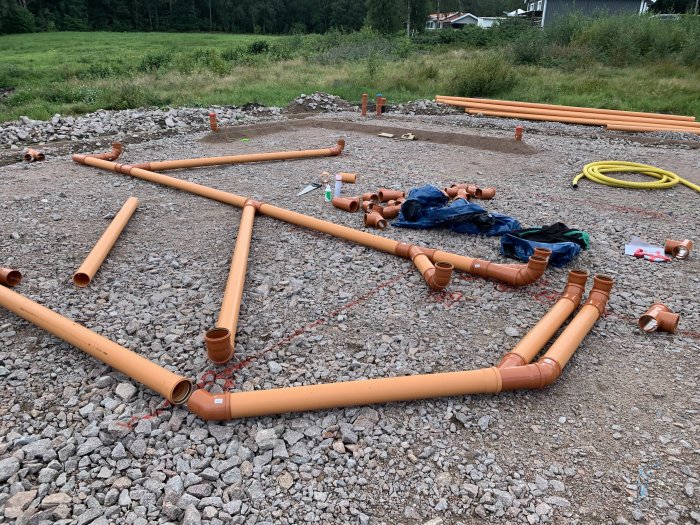 Installation av bruna avloppsrör på en grusgrund, med olika kopplingar och ledningar utspridda på marken.