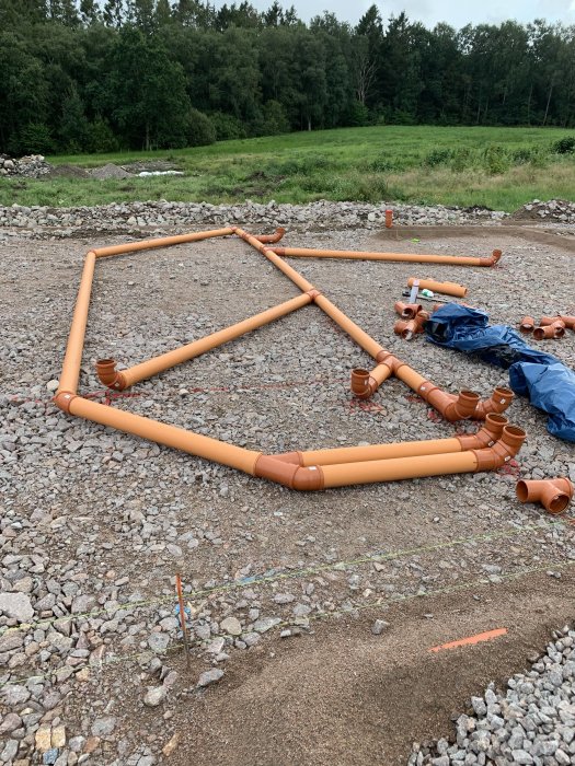 Avloppsrör och böjar i olika storlekar utspridda på grusunderlag, indikerar förberedelser för avloppsdragning i byggprojekt.