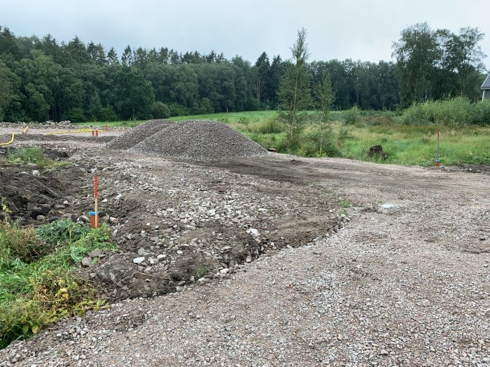 Byggplats med ny väg och garageinfart under arbete, hög med makadam och avloppsmarkeringar synliga.