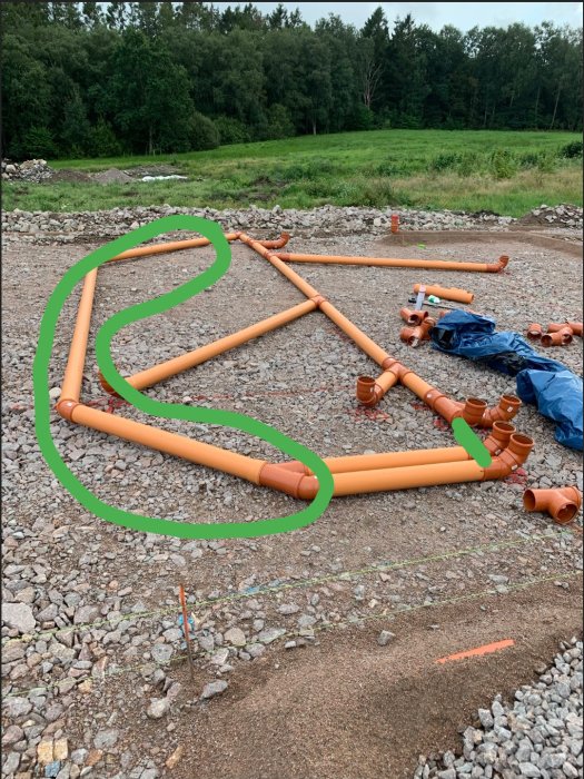 Orangea avloppsrör med lång dragning markerad i grönt på grusmark med skog i bakgrunden.