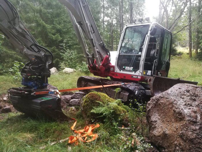 En grävmaskin fastnad i terrängen med larvbanden löstagbara, omgiven av grönska och stenar.