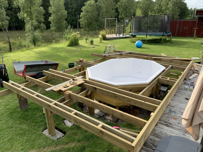 Pågående bygge av en trädterass med en vit tom swimmingpool i mitten, verktyg och byggmaterial syns.