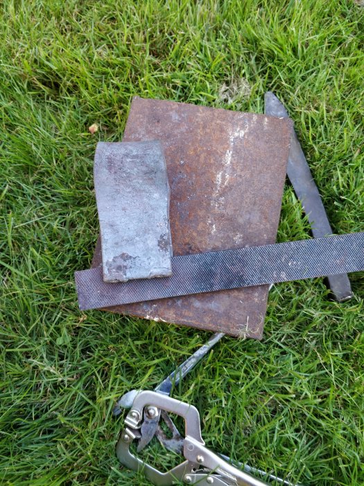 Rostiga metallbitar och stålstång på gräsmatta med gripklo, förberedelse för smide.