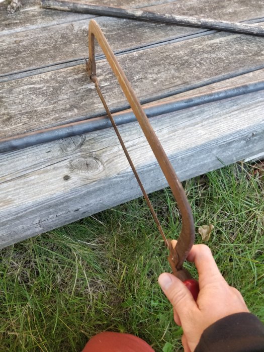 Hand som håller en hemgjord metallbåge framför träplankor och metallstång på gräs.