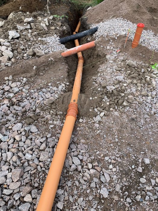 Orange vattenledningsrör med rörisolering grävt genom stenig jord med synliga dag- och dräneringsrör i bakgrunden.