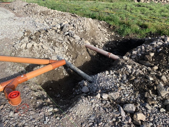 Grävd grunden för avlopp med orange avloppsrör och radonslang, samt omgivande sten och grus.