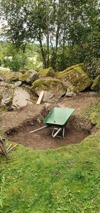 Grön skottkärra i utgrävd grop i trädgård, med stenar och verktyg runtomkring.