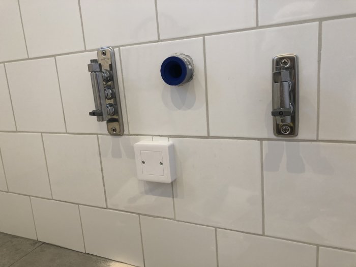 Nyinstallerade Vatette väggbrickor och badrumsfördelare på kakelvägg med synlig rörgänga och elektrisk dosa.