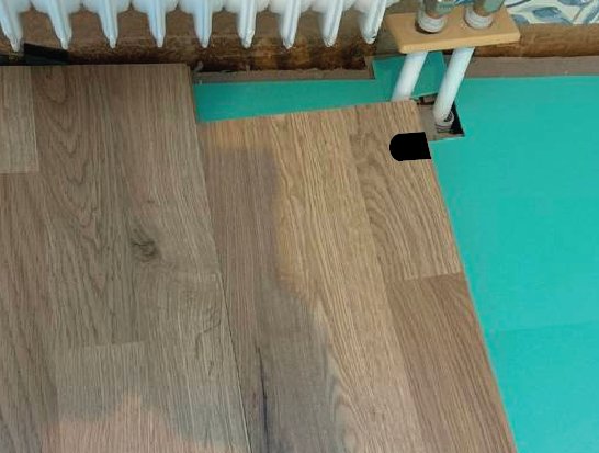 Olika golvbeläggningar med träliknande laminat intill en turkos färgad yta, nära en vit radiator.