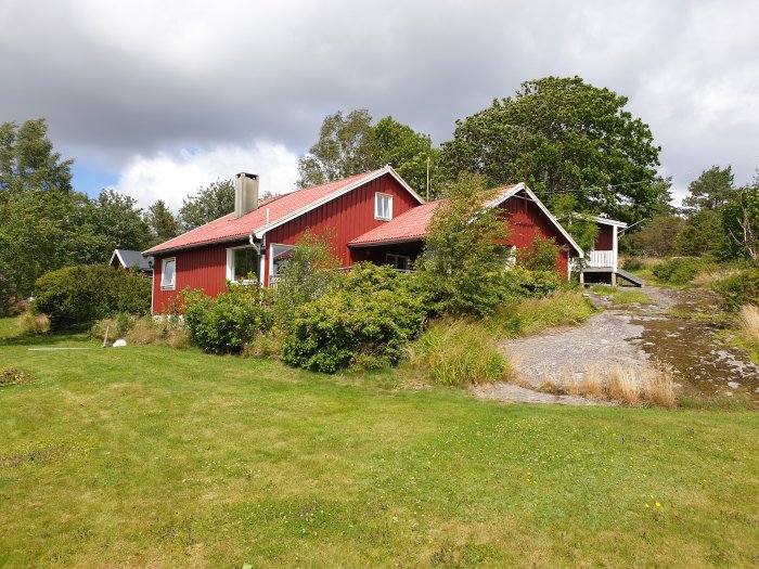 En röd 1,5-plans villa med vita knutar byggd på 1950-talet omgiven av en grönskande trädgård och bergig terräng.