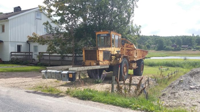 En rostig dumper parkerad på en träram ovanför postlådor med hus och sjö i bakgrunden.