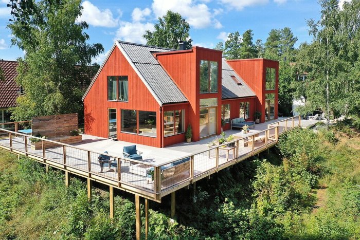 Modernt rött hus med svart tak och stora fönster på en förhöjd terrass omgiven av grönska.