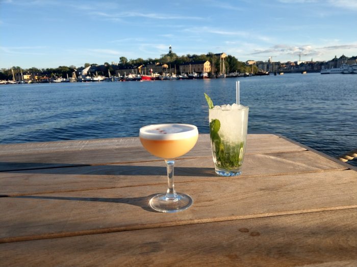 Två drinkar på ett träbord med Stockholm skyline och sjö i bakgrunden innan solnedgång.