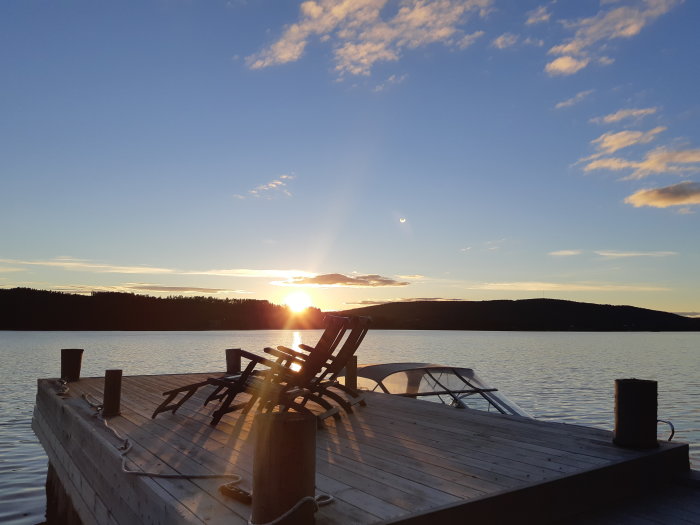 Solnedgång över en sjö med silhuett av ett bryggdäck och solstolar, lugnt vatten och himmel med moln.