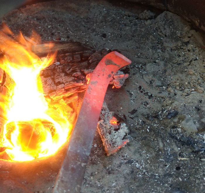 Eld i en ombyggd grop med glödande ved och en rödglödande metallstång mot lerkanten.