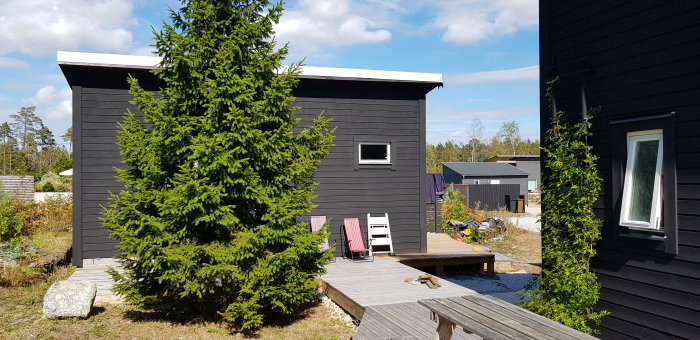 Svart trähus med fönster och altan, omgiven av träd och annan bebyggelse i bakgrunden.